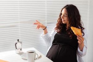 feliz embarazada joven hermosa mujer comiendo croissant y mirando a través de las persianas durante el desayuno de la mañana. concepto de mañana agradable y actitud positiva durante el embarazo foto