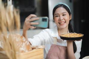 Young woman wears apron taking selfie photo with homemade pie in kitchen. Portrait of beautiful Asian female baking dessert and having fun taking photo by smartphone for online social. Home cookery.