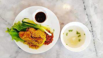 Top view of Chicken Biryani with black sweet sauce, dry onion, fresh green cabbage, cucumber, spring onions on white dish and soup on bowl on marble table with copy space. Flat lay of Asian food. photo