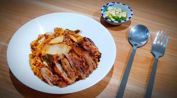 Kimchi fried rice with grilled pork on white dish with spoon and fork on wooden background. Asian food with chili, garlic in fish sauce on wood table photo