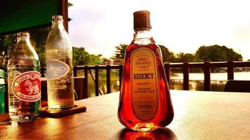 Bangkok, Thailand- January 1, 2023 Thai whisky, soda and water on brown table with sunlight of sunset and lake, tree and sky background. Refresh drinking, Relaxing time with friend and Join party photo
