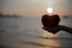 fondo borroso de forma de corazón rojo a mano en la costa con puesta de sol de silueta en el mar para crear una tarjeta dulce y un afiche de pareja. foto