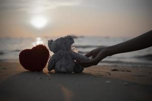 female hand holding small lonely purple cute little bear sitting with red handicraft heart on the beach for created postcard  of international missing children, broken heart. photo
