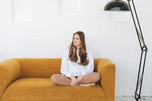 Young woman teenager in white hoodie beautiful girl sitting on yellow couch in the modern interior photo