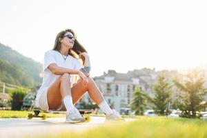 mujer joven delgada con cabello largo y rubio con ropa deportiva ligera sentada con longboard en el parque de patinaje al aire libre al atardecer foto