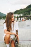 Slim young woman with long blonde hair in light sports clothes sitting with longboard in the outdoor skatepark at sunset photo