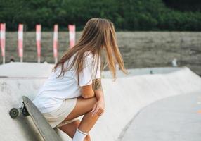 Slim young woman with long blonde hair in light sports clothes with longboard in outdoor skatepark photo