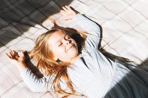 Una linda y graciosa niña con el pelo largo vestido de casa gris divirtiéndose yace a la luz del sol en la cama en casa foto