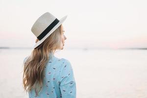 Beautiful blonde young woman in blue dress and straw hat on pier on sunset photo