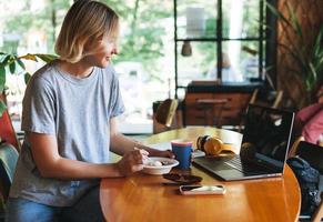 joven mujer rubia sonriente independiente con auriculares amarillos trabajando en un cuaderno en la mesa del café. niña estudiante niña estudiando en un curso en línea. mujer desayunando en la cafetería foto