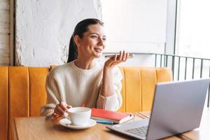 Attractive young brunette smiling woman student freelancer with cup of tea using mobile phone and laptop working in cafe photo