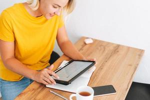 Young blonde woman in yellow t-shirt studies cryptocurrency charts and stock quotes on digital tablet with cup of tea in kitchen at home photo