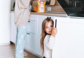 Cute little girl with long hair looks out from behind door in kitchen at home. Daughter helps mother with the cooking of food photo