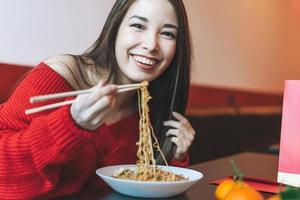 bella joven asiática sonriente vestida de rojo comiendo comida asiática con palillos de bambú en el restaurante vietnamita chino foto