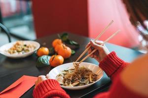 bella joven asiática sonriente vestida de rojo con un amigo comiendo fideos con palillos de bambú en el restaurante vietnamita chino foto