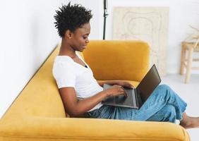 Beautiful young African American woman in white t-shirt and blue jeans using laptop sitting on yellow sofa in bright modern interior photo