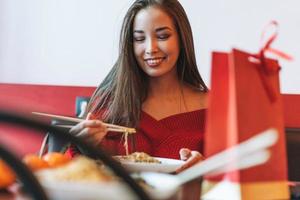 Beautiful smiling young asian woman in red clothes eating noodles with bamboo chopsticks in the chinese vietnamese restaurant photo