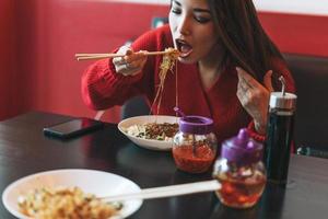 bella joven asiática sonriente vestida de rojo comiendo fideos con palillos de bambú en el restaurante vietnamita chino foto