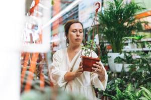 una mujer morena de mediana edad con vestido blanco compra plantas de interior en macetas verdes en la tienda del jardín foto