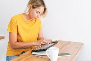 Una joven rubia con camiseta amarilla estudia gráficos de criptomonedas y cotizaciones de acciones en una tableta digital con una taza de té en la cocina de casa foto