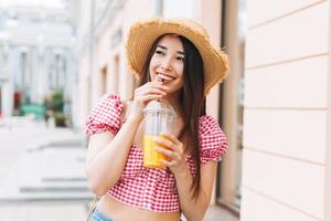 sonriente hermosa joven asiática con el pelo largo en sombrero de paja bebiendo jugo de naranja en la calle de la ciudad foto