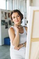 Thinking charming young woman artist in white casual clothes with paint brushes in front of easel in the modern art gallery photo