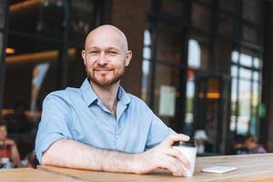 Adult bald smiling attractive man forty years with beard in blue shirt businessman using mobile phone with paper cup of coffee at cafe photo