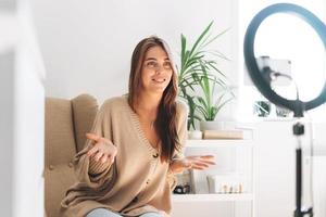 joven bloguera de mujer bonita en cardigan acogedor graba video en el teléfono móvil usando una lámpara de anillo sentada en una silla en el hogar foto