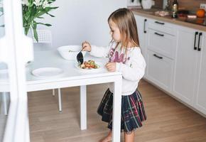 Pretty little girl with long hair puts on vegetable salad in kitchen with bright interior at home photo