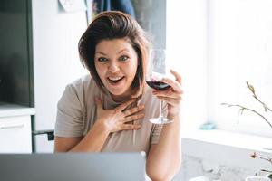 Beautiful happy emotional brunette young woman plus size body positive with glass of wine having video call on laptop at home photo