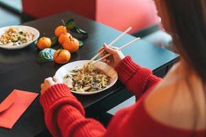 Beautiful smiling young asian woman in red clothes eating noodles with bamboo chopsticks in chinese vietnamese restaurant photo