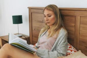 amable y encantadora mujer rubia leyendo un libro en la cama en un interior luminoso en casa foto