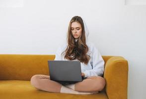 Young woman in white hoodie beautiful girl freelancer student using laptop sitting on yellow couch in modern interior photo