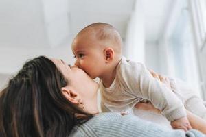 joven madre besando a su lindo bebé en las manos en una habitación luminosa emoción de amor foto