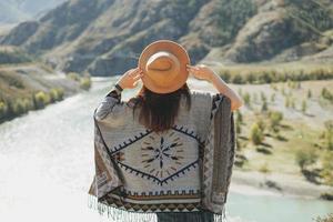 Carefree brunette young woman traveler in poncho from back on background of the mountain river photo