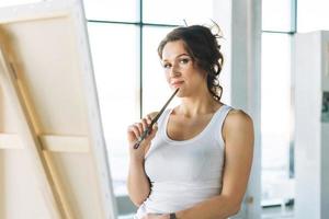 Thinking charming young woman artist in casual clothes with paint brushes in front of easel in modern art gallery photo