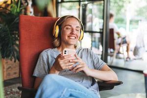 joven mujer rubia sonriente con los ojos cerrados en auriculares amarillos disfruta de la música con el teléfono móvil sentado en una silla en el café foto