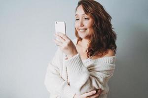 Attractive friendly smiling brunette woman middle age in cozy knitted cardigan using mobile isolated on grey background photo