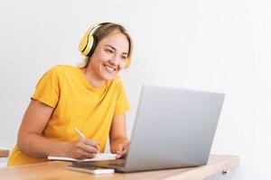 joven mujer rubia sonriente en camiseta amarilla con auriculares trabajando en la computadora portátil en la cocina en casa foto