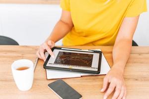 Una joven rubia con camiseta amarilla estudia gráficos de criptomonedas y cotizaciones de acciones en una tableta digital con una taza de té en la cocina de casa foto