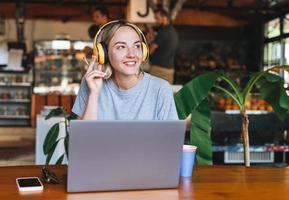 Young smiling blonde woman freelancer in yellow headphones working on laptop on table at cafe photo