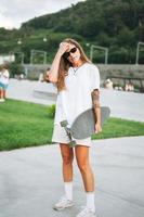 Slim young woman with long blonde hair in light sports clothes with longboard in the outdoor skatepark at sunset photo