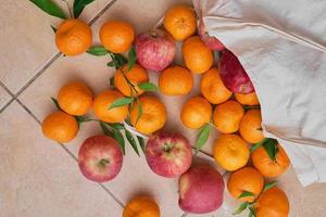 Colorful ripe fruits in a reusable eco bag on a tiled floor. Delivery of tangerines and apples. Healthy food, vitamins. View from above. photo