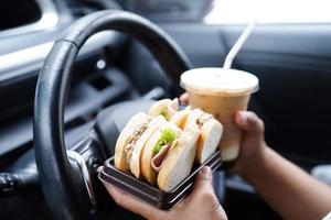 Asian woman driver hold ice coffee cup and sandwich bread for eat and drink in car, dangerous and risk an accident. photo