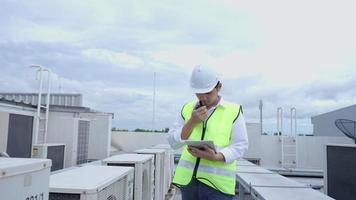 un ingénieur de maintenance asiatique utilise un talkie-walkie pour expliquer les travaux sur le toit de l'usine. l'entrepreneur inspecte le système de compresseur et planifie l'installation de systèmes de climatisation sur le chantier de construction video