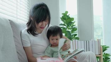 heureuse mère asiatique se détendre et lire un livre avec bébé temps ensemble à la maison. un parent s'assoit sur un canapé avec sa fille et lit une histoire. apprendre le développement, la garde d'enfants, le rire, l'éducation, la narration, la pratique. video