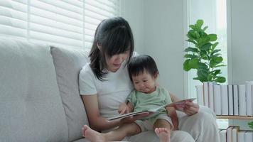 feliz mãe asiática relaxa e lê o livro com o tempo do bebê juntos em casa. pai senta no sofá com a filha e lê uma história. aprender desenvolvimento, cuidar de crianças, rir, educação, contar histórias, praticar. video