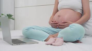 schwangere frau, die mit ihrem ehemann eine videokonferenz nimmt. eine schwangere frau winkt ihrem mann zu und zeigt die für ihre tochter vorbereiteten rosa schuhe. Fernkommunikationskonzept. video