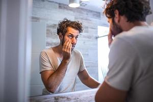 Young caucasian man taking care of his face looking at mirror reflection in bathroom morning clean eyes. Reflection of handsome man with beard looking at mirror and touching face in bathroom grooming photo