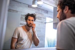 higiene matutina, hombre guapo en el baño mirándose en el espejo. reflejo de un hombre guapo con barba mirando al espejo y tocando la cara en el aseo del baño foto